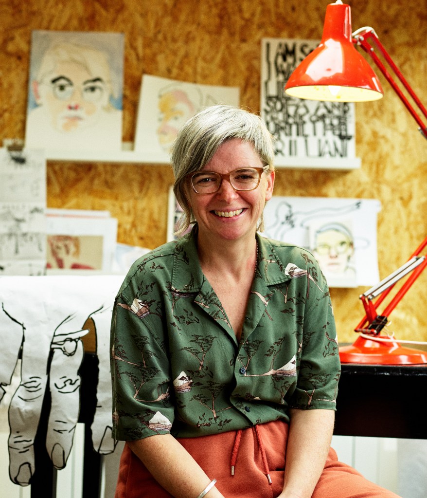 Portrait photograph of Alys in her studio, smiling at the camera. She is a white woman with short grey hair and glasses, wearing  green shirt. Drawings can be seen behind her.
