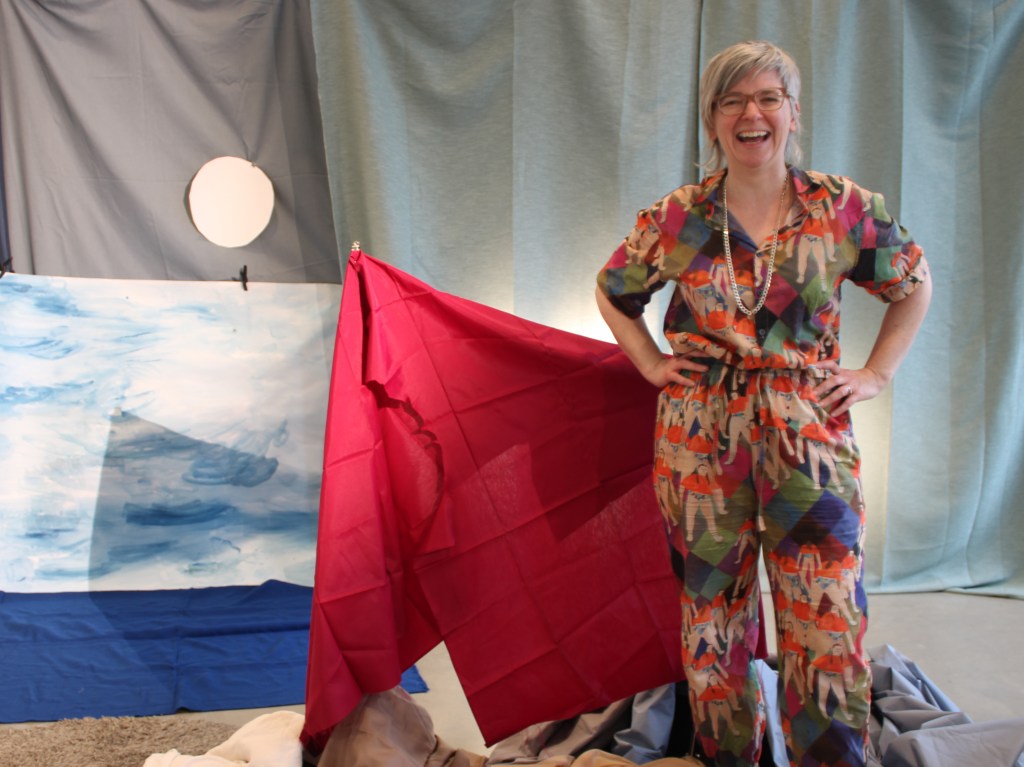 Alys, a white woman with grey hair and glasses, looks towards the camera, laughing. She is standing in front of a fabric and paper landscape.