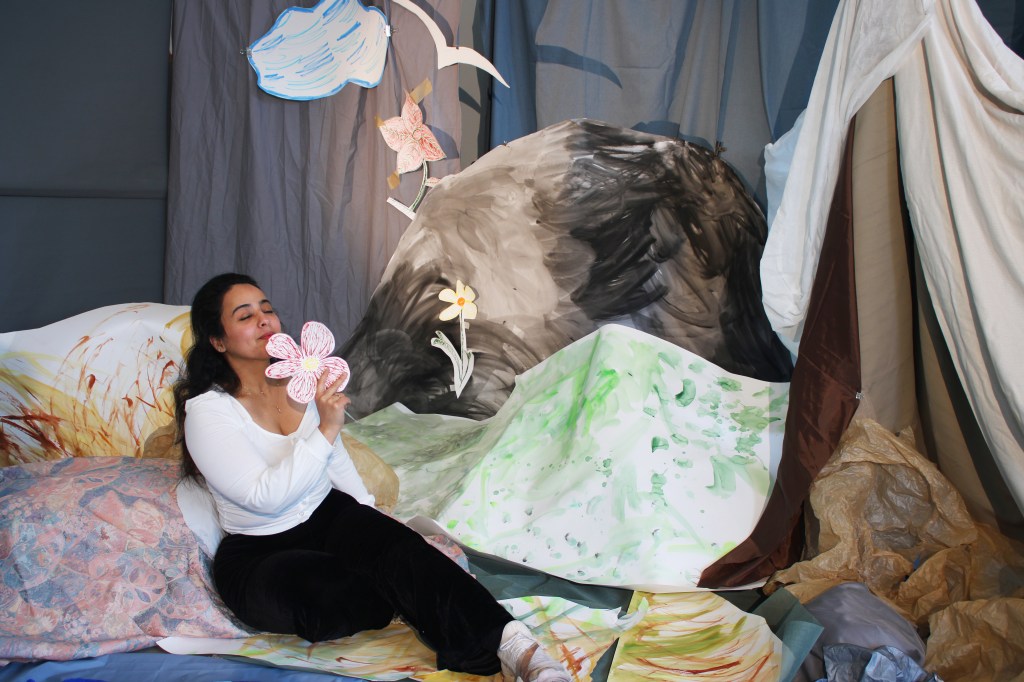 A brown skinned woman with long black hair, sits amidst a mountainous landscape made of paper and fabric. Her eyes are closed and she is smelling a paper flower.